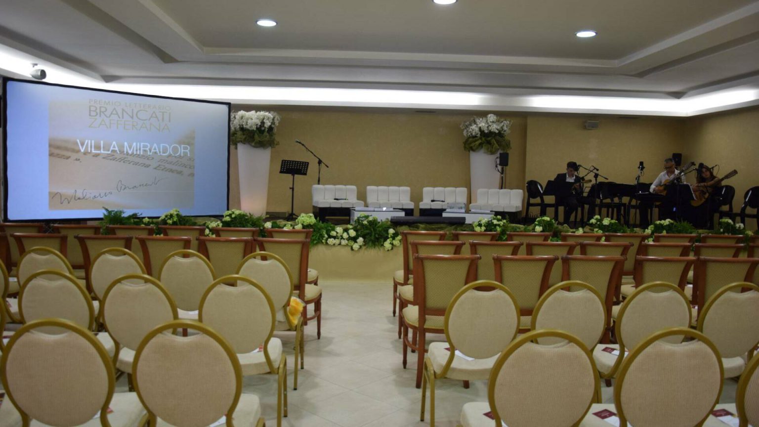 Sala Congressi: file di sedie rivolte verso il tavolo dei relatori e lo schermo