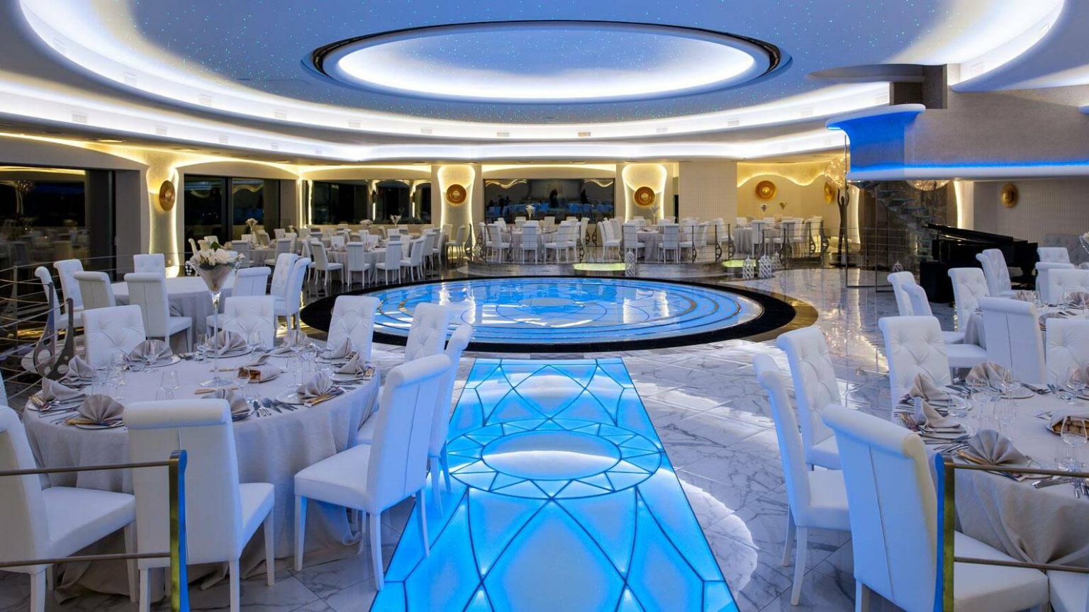 Sala Gloria: tavoli bianchi attorno a una passerella luminosa, sotto un soffitto a cerchi di luce